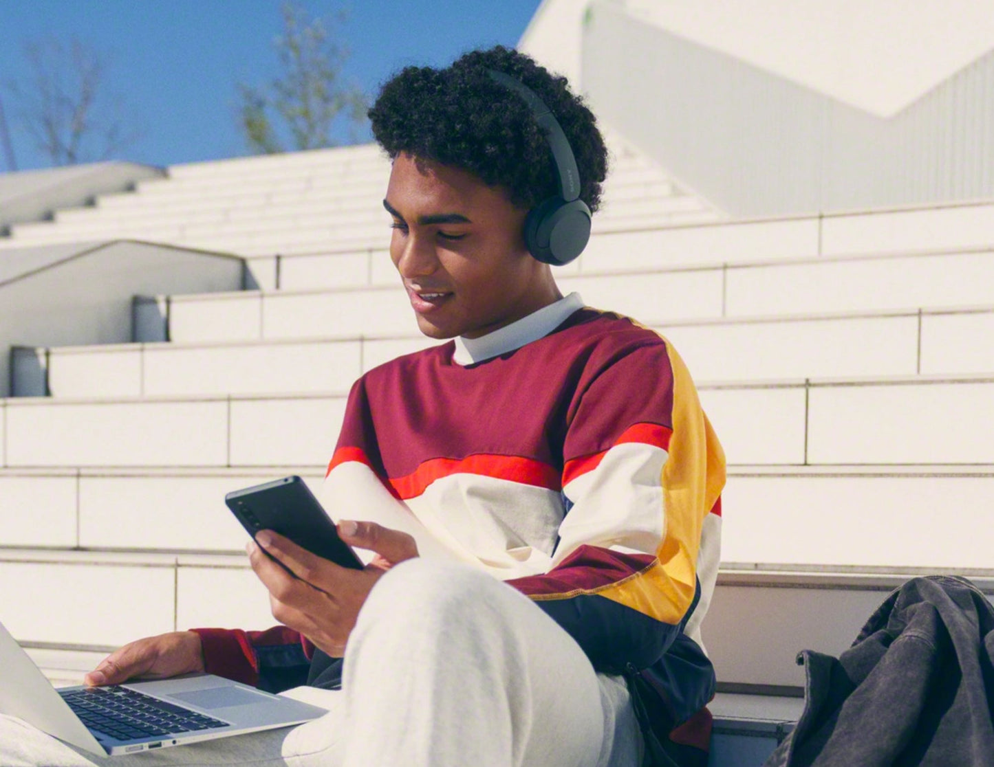 Person sitting outdoors using a laptop and smartphone, wearing headphones.