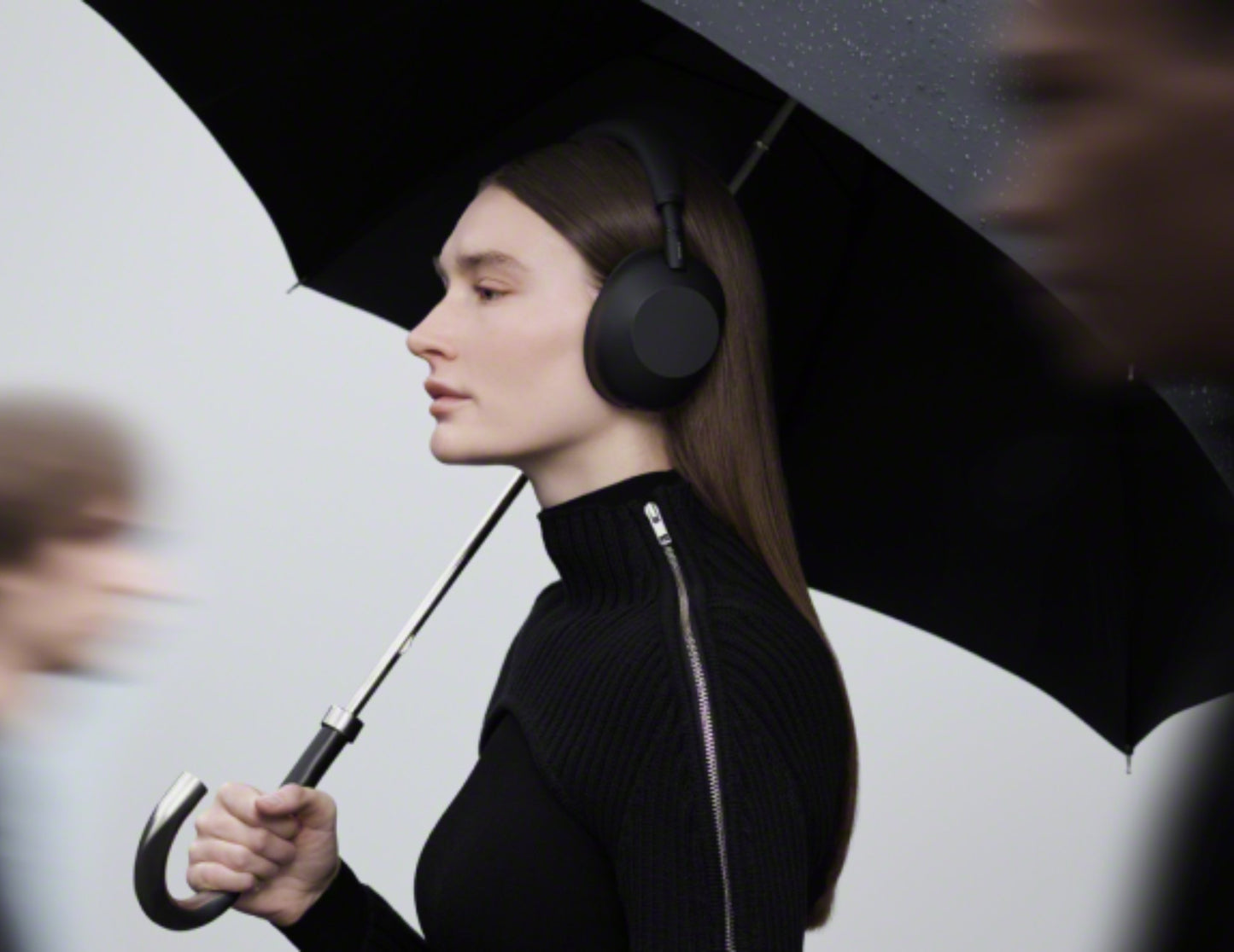 Woman wearing Sony WH-1000XM6 Wireless Noise Canceling Headphones – Black holding a black umbrella against a white background