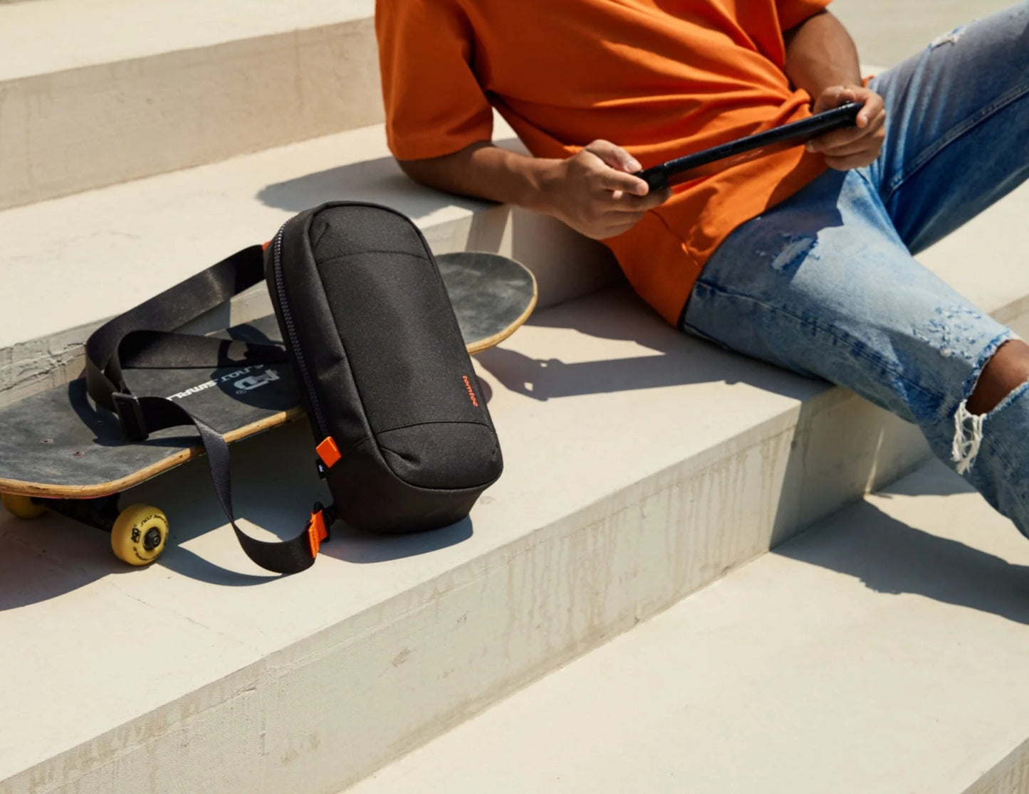 Person sitting on steps with a skateboard and Tomtoc GameOn-G49 Nintendo Switch 2 Sling Bag – Black (G49S1D1), wearing an orange shirt and blue jeans.