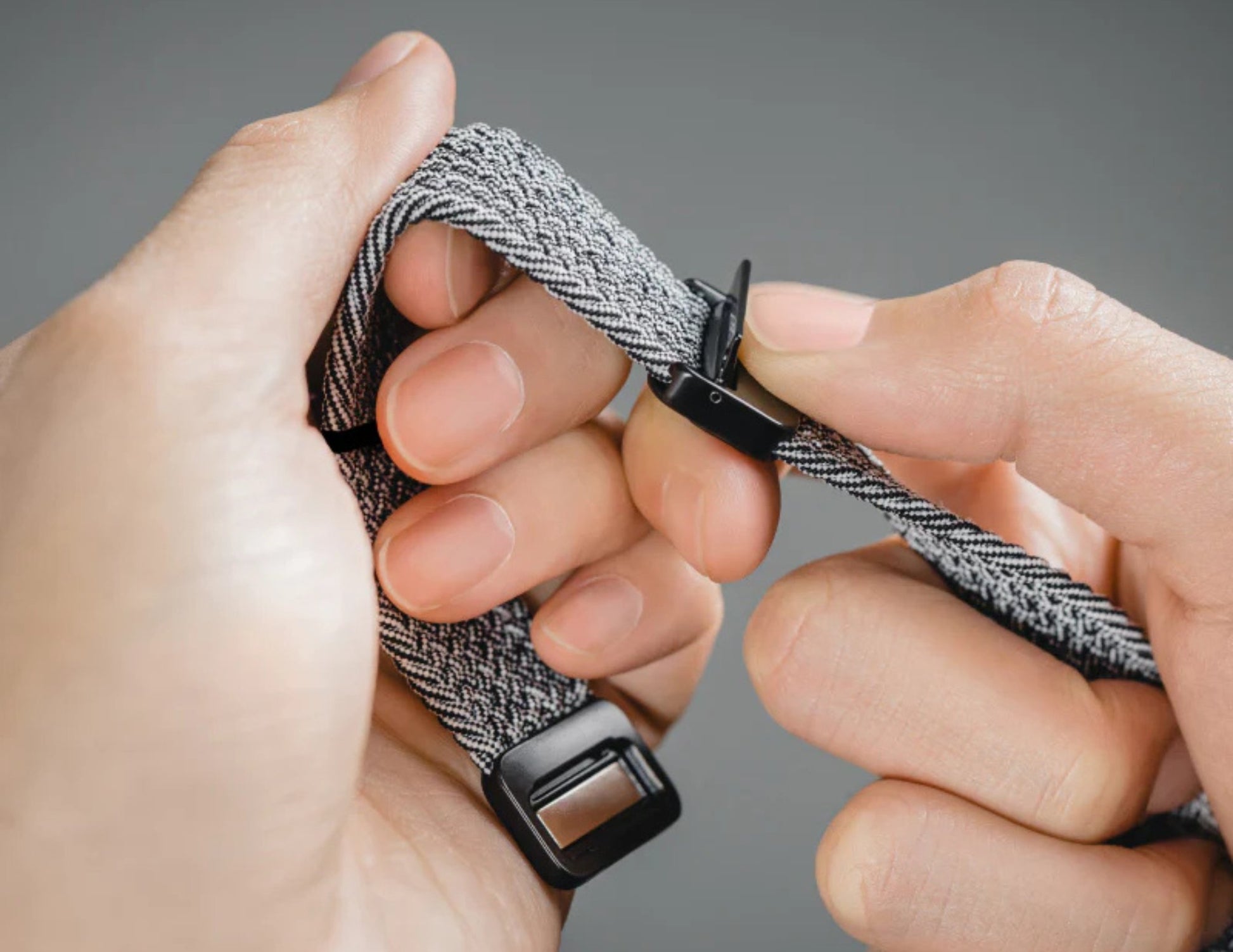 Close-up of hands adjusting a gray and black woven bracelet on a plain background.