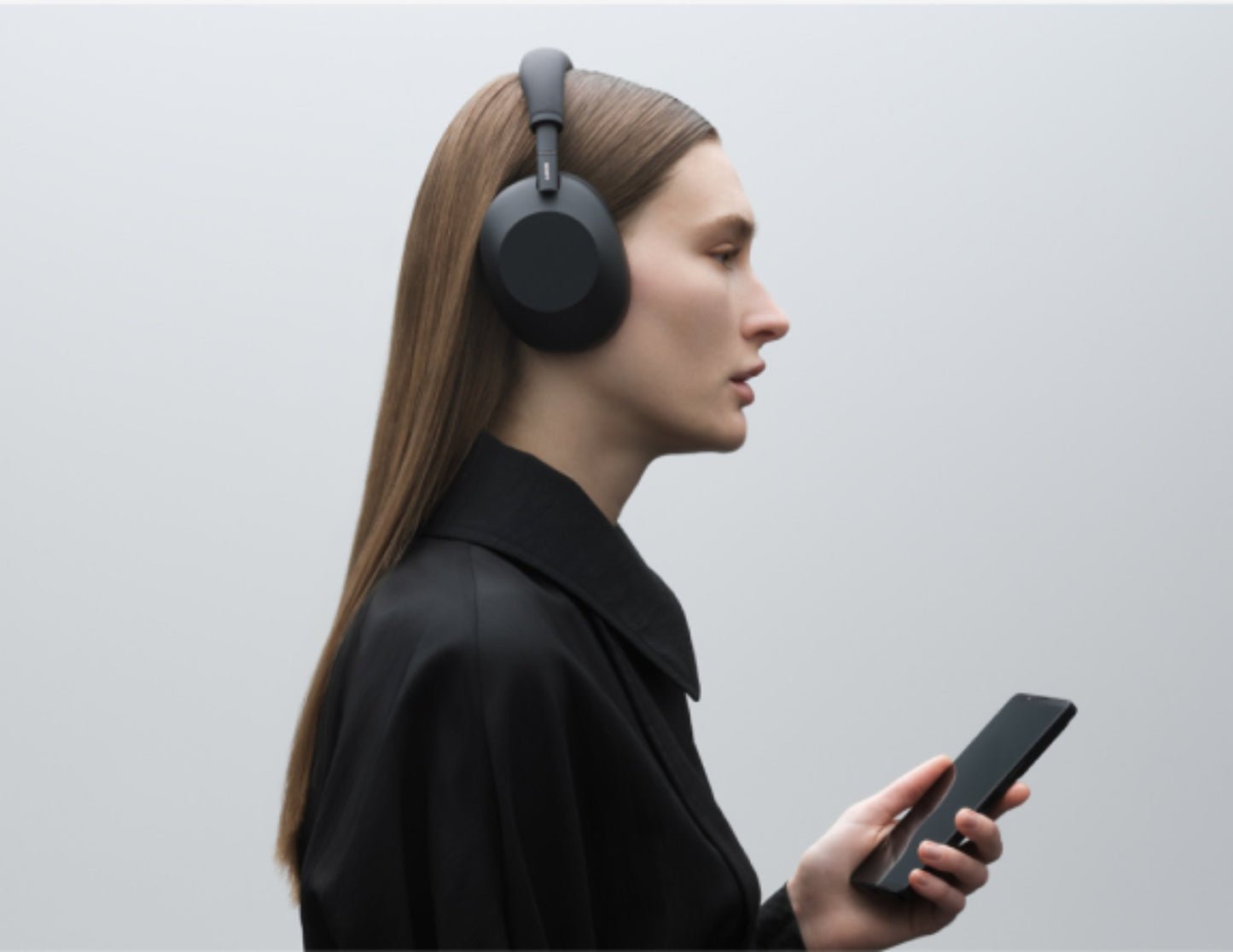 Person wearing Sony WH-1000XM6 Wireless Noise Canceling Headphones – Black and holding a phone against a light gray background