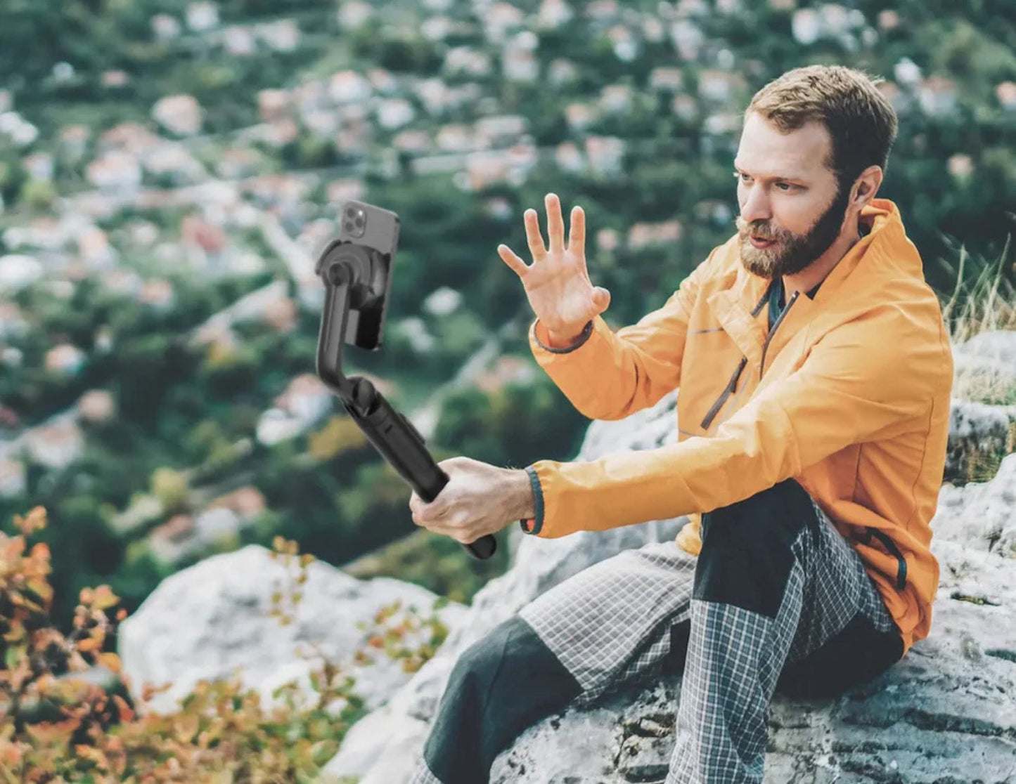 Man in an orange jacket using a Momax 1-Snap Pole Magnetic Tripod Selfie Stick with Wireless Remote – KM17D in a mountainous area