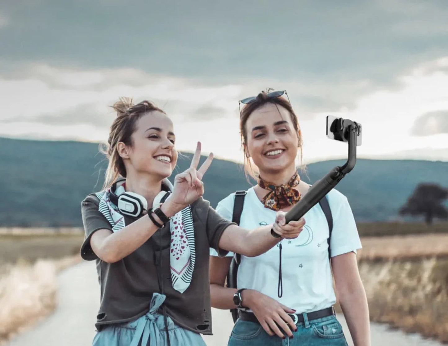 Two women outdoors, one holding a Momax 1-Snap Pole Magnetic Tripod Selfie Stick with Wireless Remote – KM17D, with a scenic background.