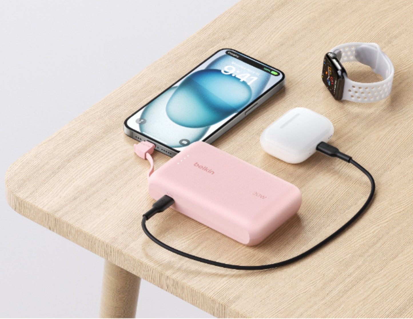 Pink Belkin BoostCharge BPB021hq 10K 20W power bank with USB-C cable from Thabthaba Store with white earbuds, and smartwatch on a wooden table.