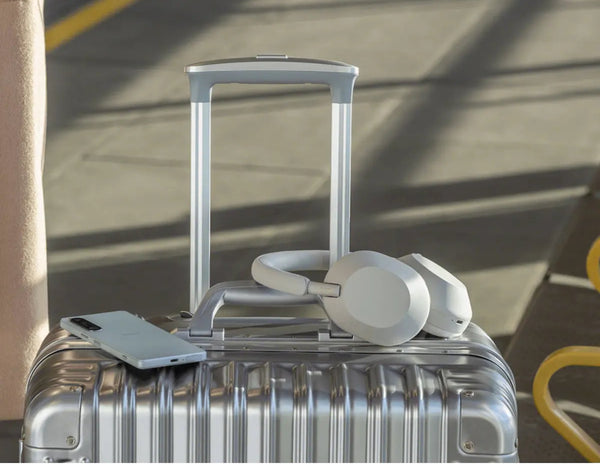 Silver suitcase with a phone, white earbuds, and a white charging cable on top, against a blurred background.