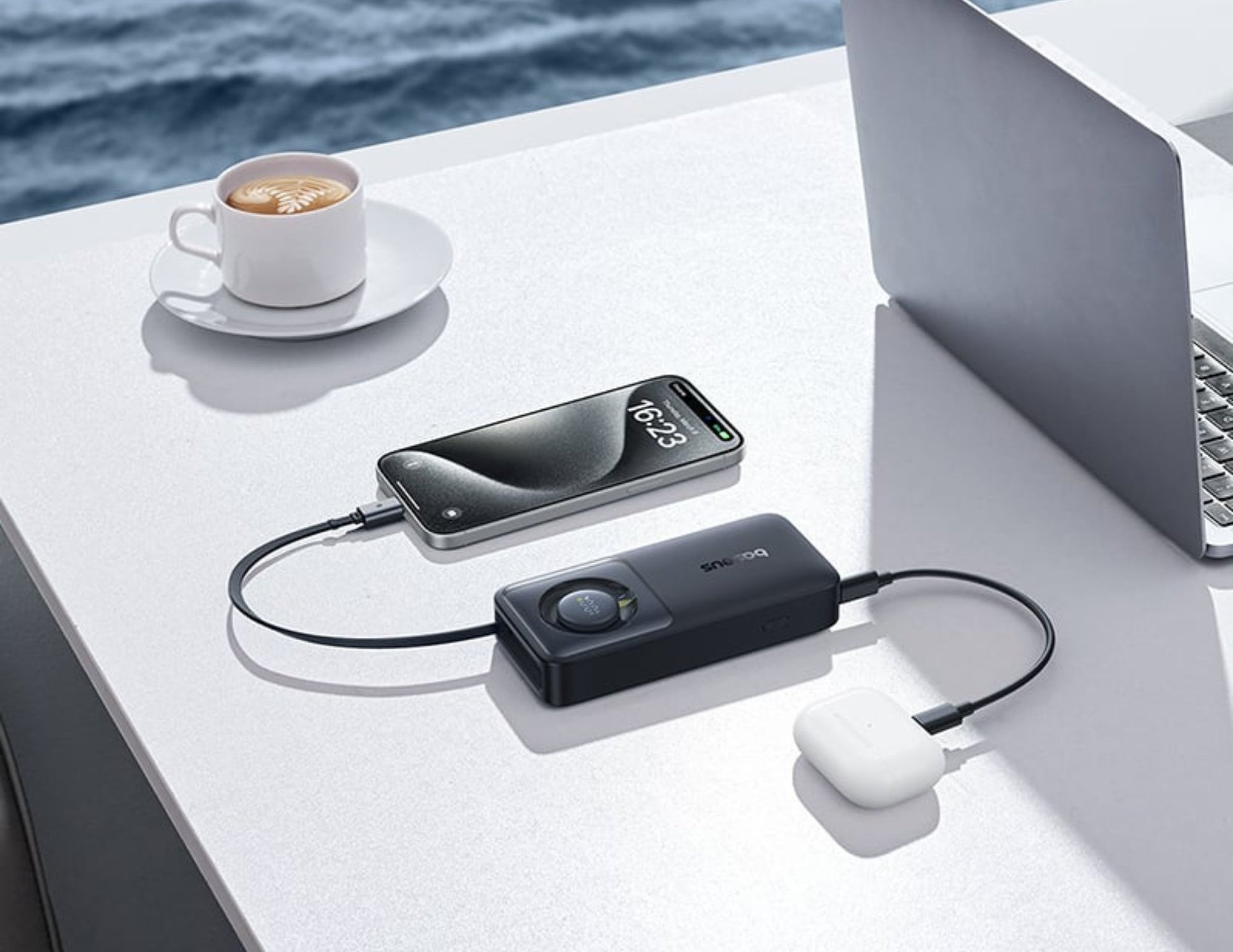 Laptop, smartphone, and power bank on a table with a cup of coffee and scenic background.