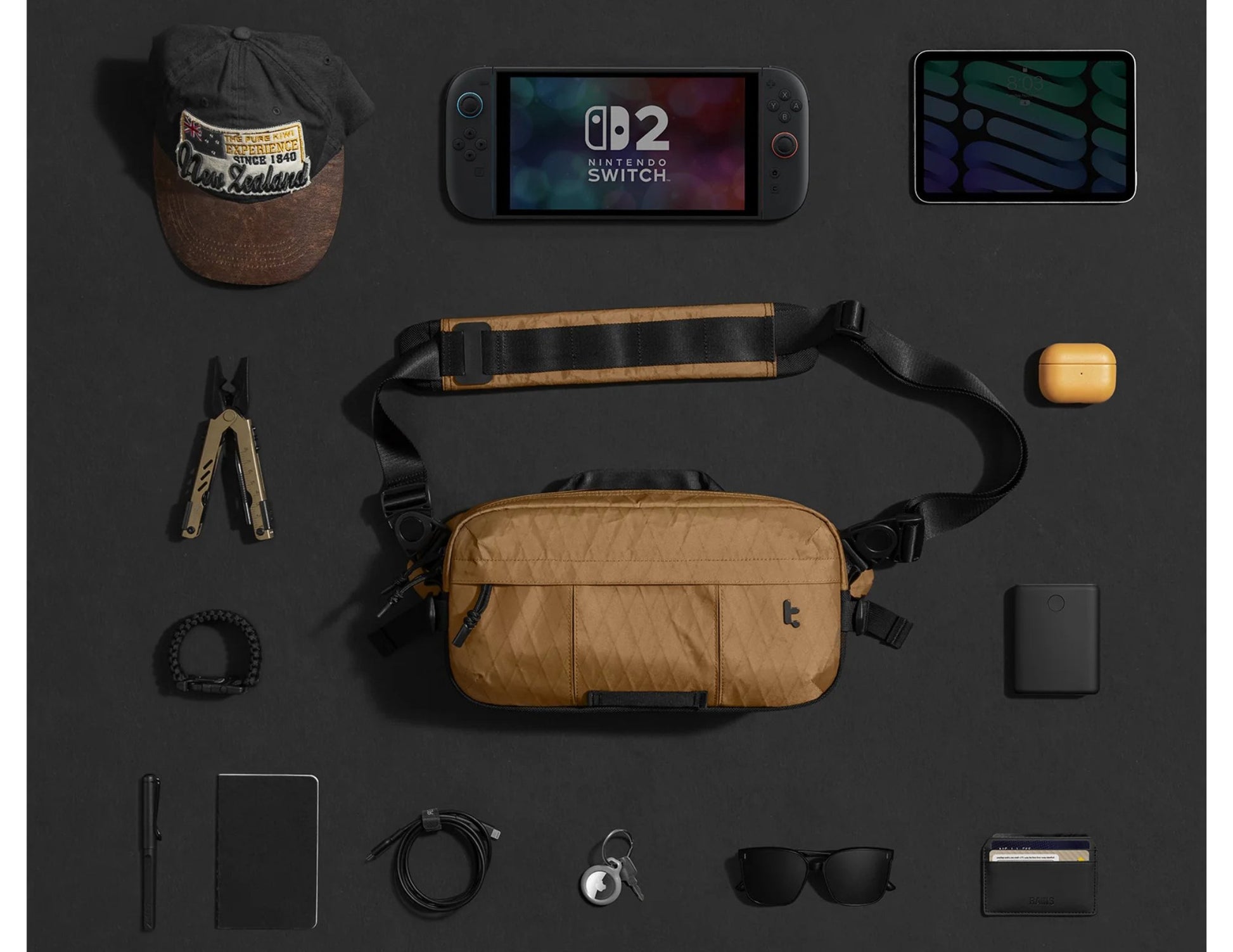 Collection of items including a Nintendo Switch console,Tomtoc Wander-T26 X-Pac Daily Sling Bag – Brown (T26S1K1), and various accessories on a dark surface.