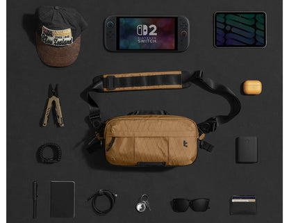 Collection of items including a Nintendo Switch console,Tomtoc Wander-T26 X-Pac Daily Sling Bag – Brown (T26S1K1), and various accessories on a dark surface.