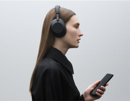 Person wearing Sony WH-1000XM6 Wireless Noise Canceling Headphones – Black and holding a phone against a light gray background