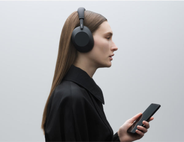 Person wearing Sony WH-1000XM6 Wireless Noise Canceling Headphones – Black and holding a phone against a light gray background