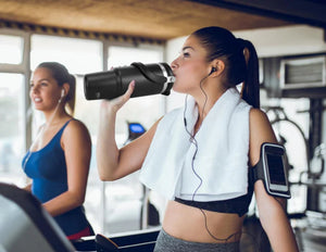 Woman in a gym drinking from a Tyeso TS-8828 Vacuum Insulated Tumbler 900ML with another person in the background.