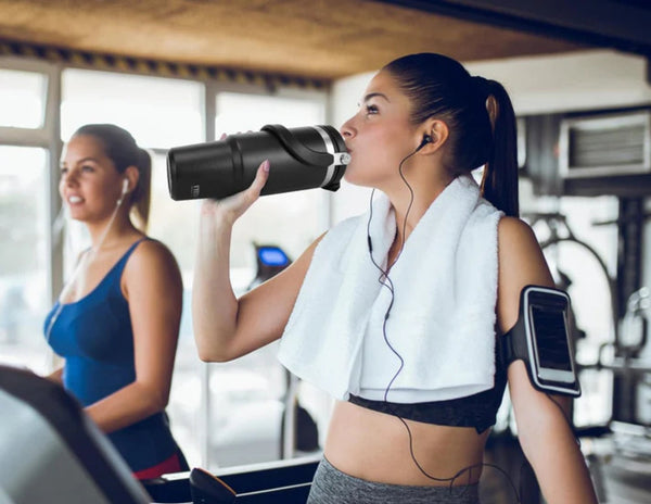 Woman in a gym drinking from a Tyeso TS-8828 Vacuum Insulated Tumbler 900ML with another person in the background.