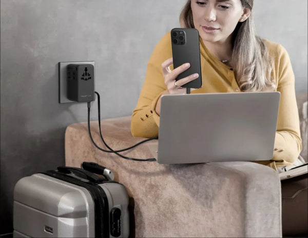 Woman using a laptop and phone with a power adapter on a couch.