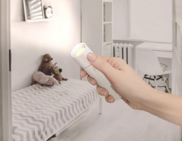 Hand holding a white remote control in a bedroom setting with a bed and stuffed animals.