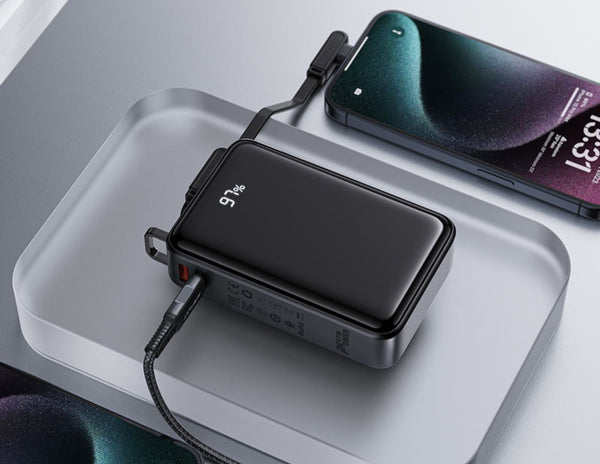 Portable power bank with charging cable on a white surface, with a smartphone in the background.