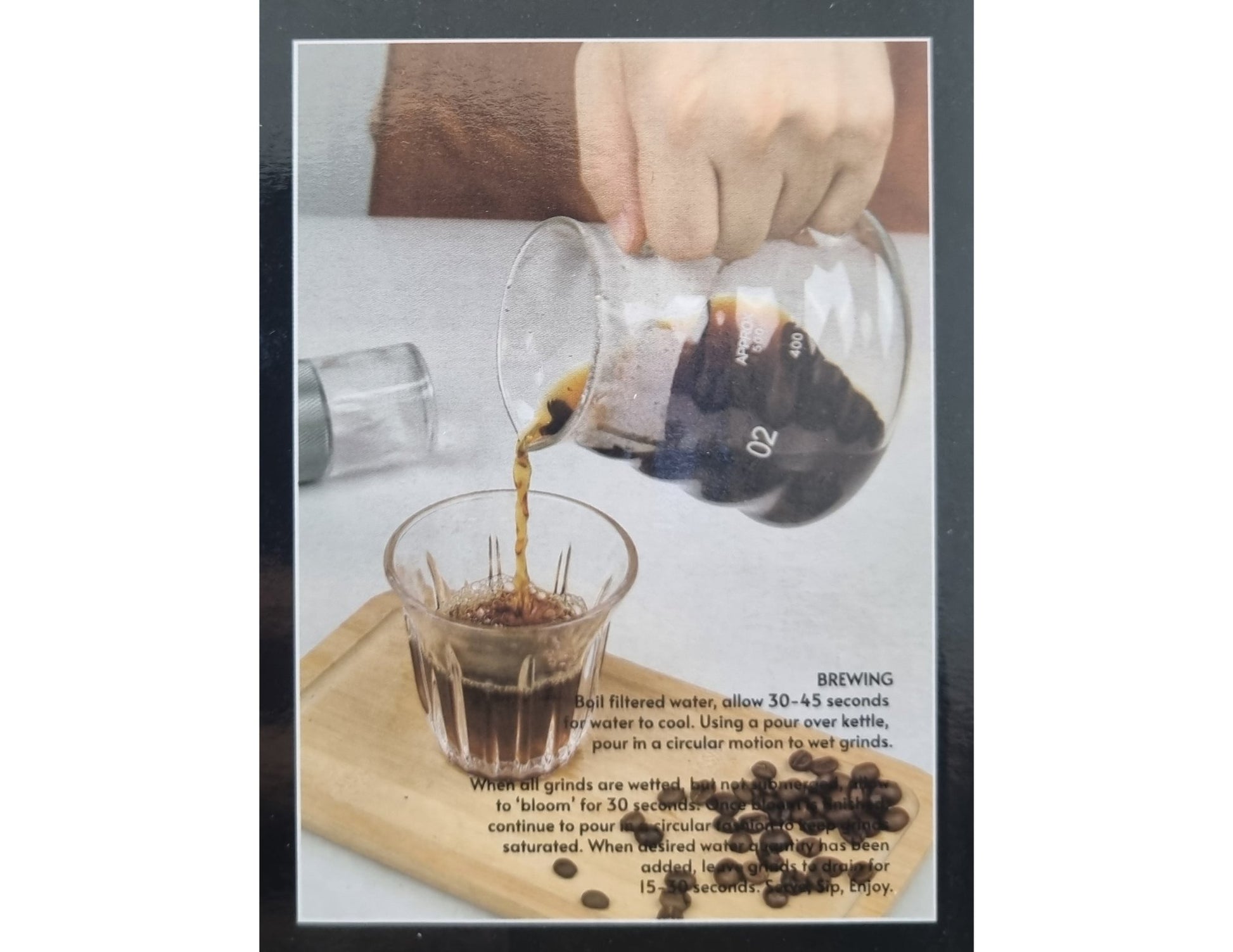 Pouring coffee from a glass container into a glass with coffee beans and brewing instructions on a wooden board.