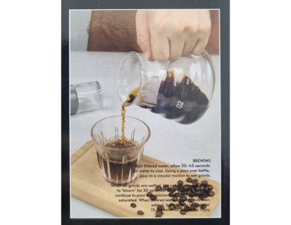 Pouring coffee from a glass container into a glass with coffee beans and brewing instructions on a wooden board.